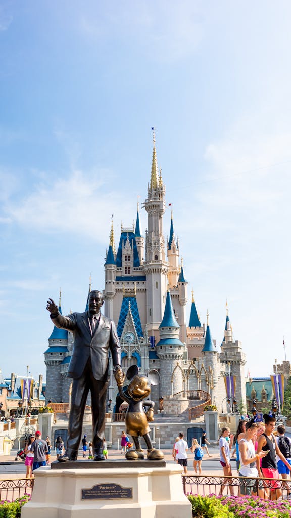 A sunny day at a famous theme park with a castle and bronze statues surrounded by tourists.